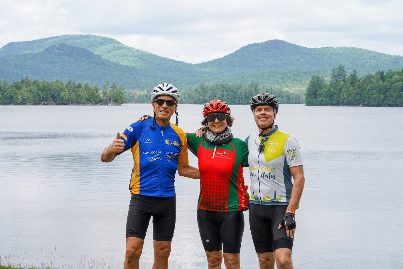 Three cyclists in helmets and cycling jerseys standing together beside a lake, with forested mountains in the background.