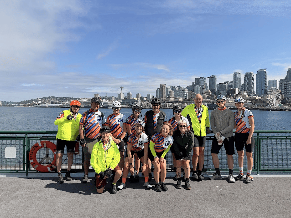 Bike the US for MS cycling team posing together on a ferry, with the Seattle skyline and Space Needle across the water in the background.