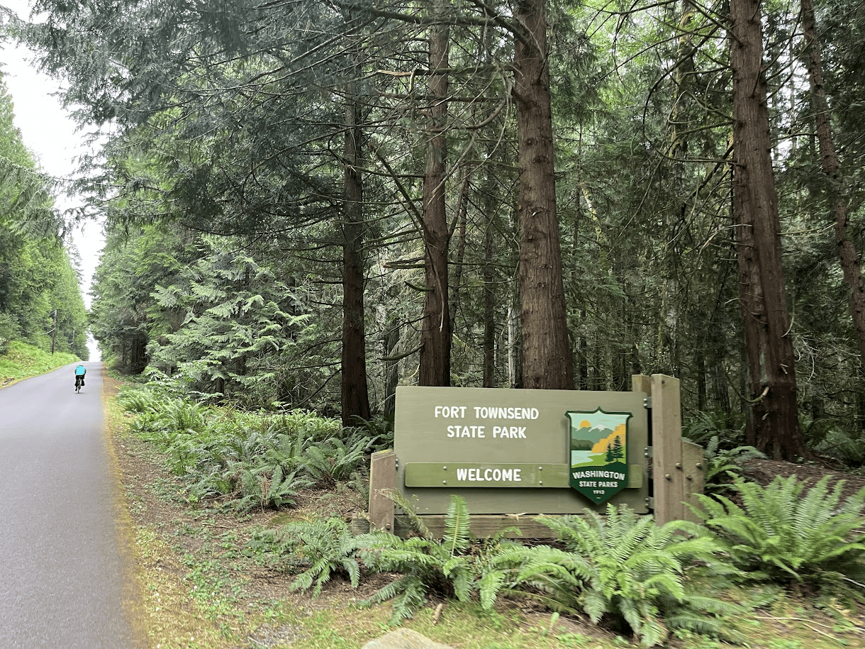 Cyclist riding along a paved road through dense forest beside the Fort Townsend State Park welcome sign in Washington.