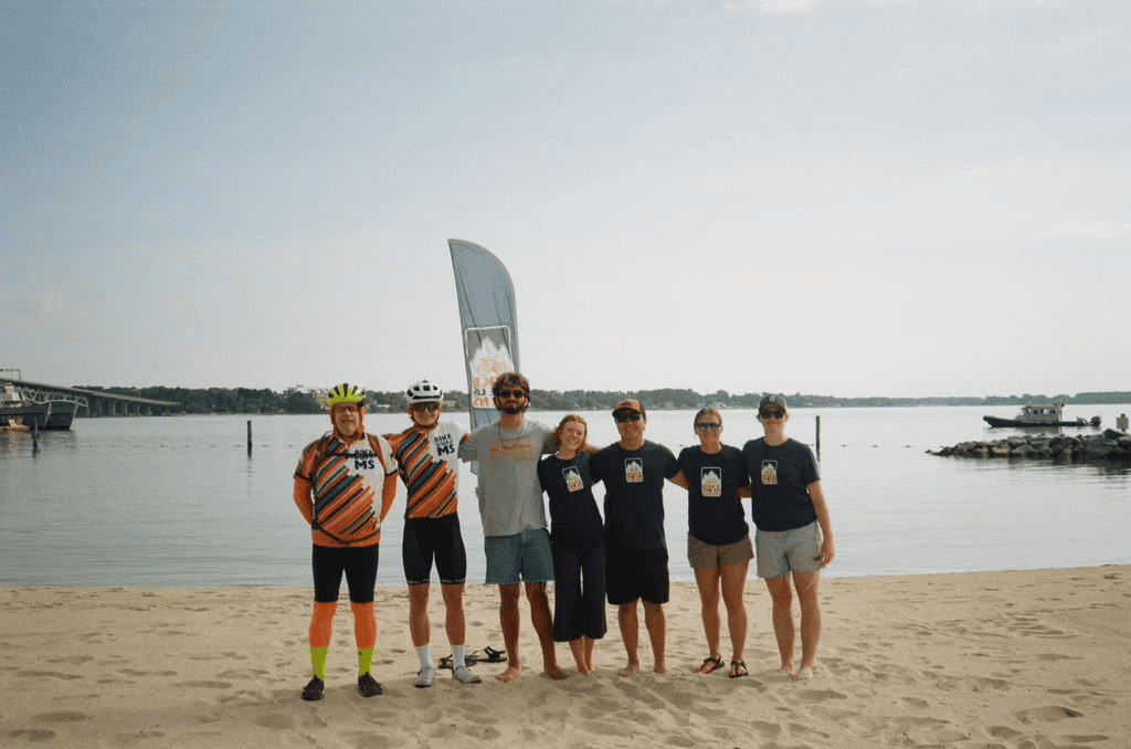 Bike the US for MS riders and route leaders standing together on a sandy beach beside the water.