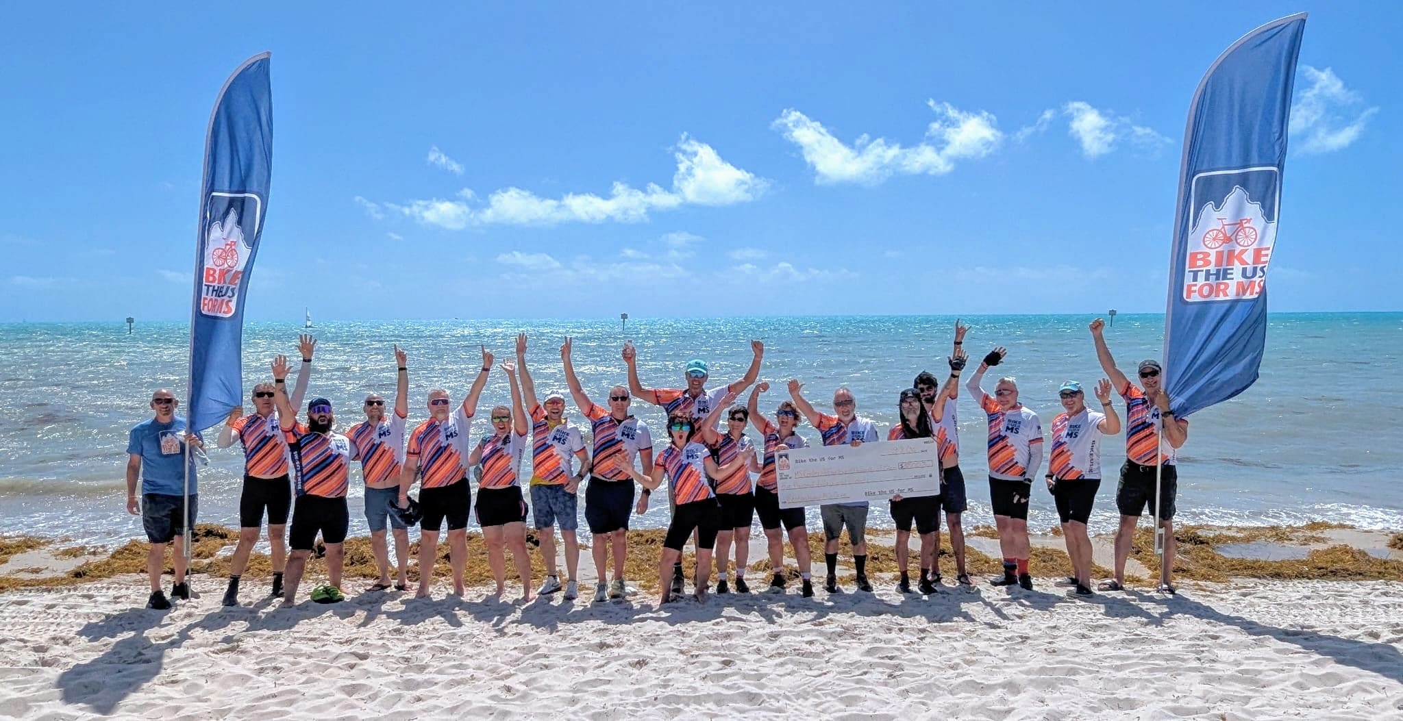 25 Bike the US for MS cyclists celebrating a check donation on the beach with a large check and Bike the US for MS flags