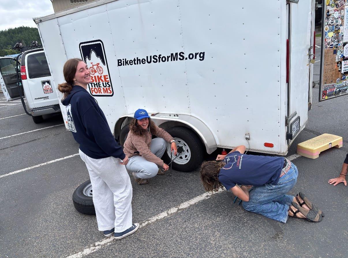 three route leaders smiling while fixing a tyre on the Bike the US for MS trailer. one  person is standing, one person is knelt tightening the bolts on the tyre, one person is knelt by the jack
