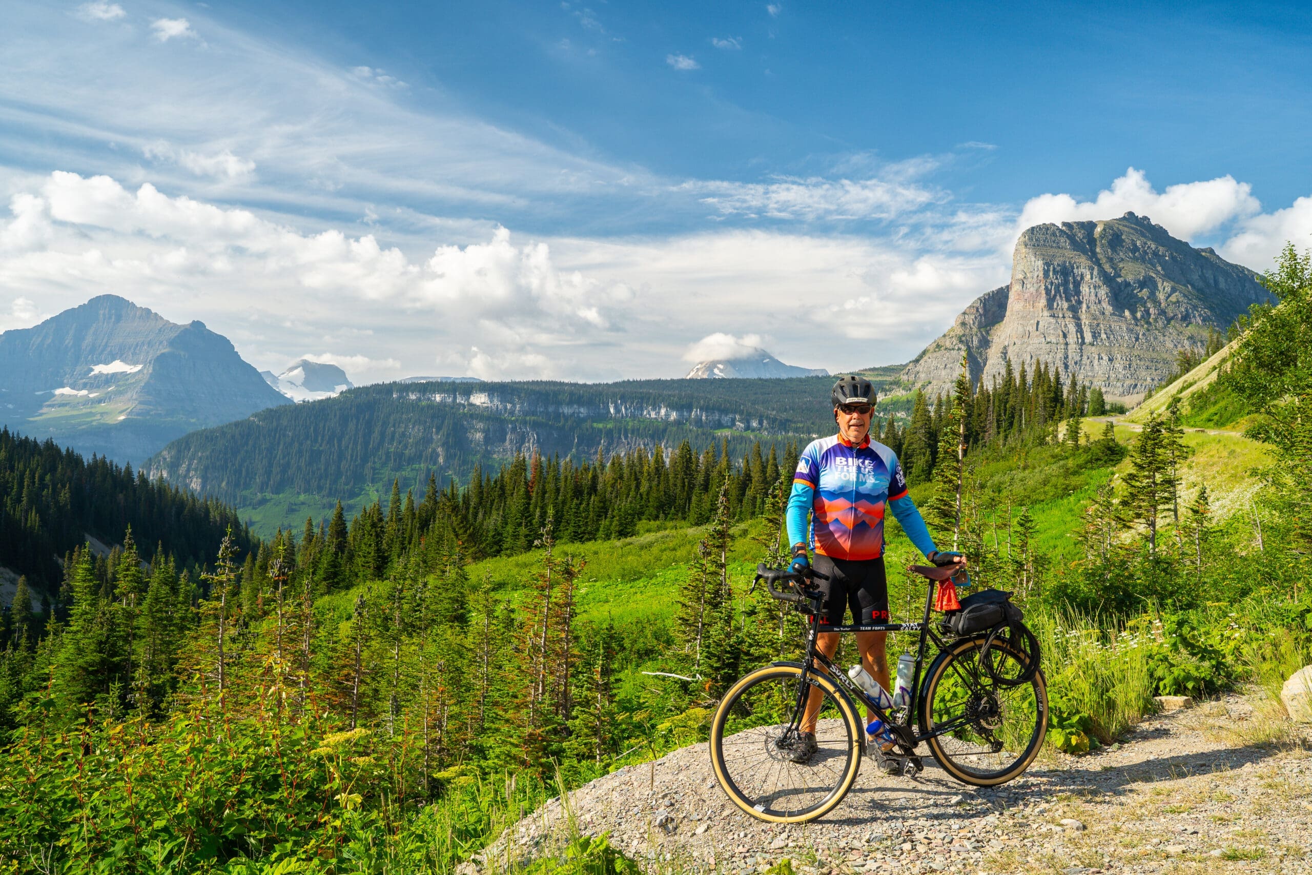 Bike the US cyclist standing with his bike with Glacier National Park mountain views behind him.